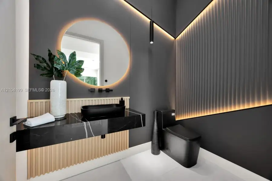 Floating vanity with stone countertop and matte black vessel sink, circular backlit mirror, vertical slat wood accent paneling, integrated recessed cove lighting, and matte black plumbing fixtures