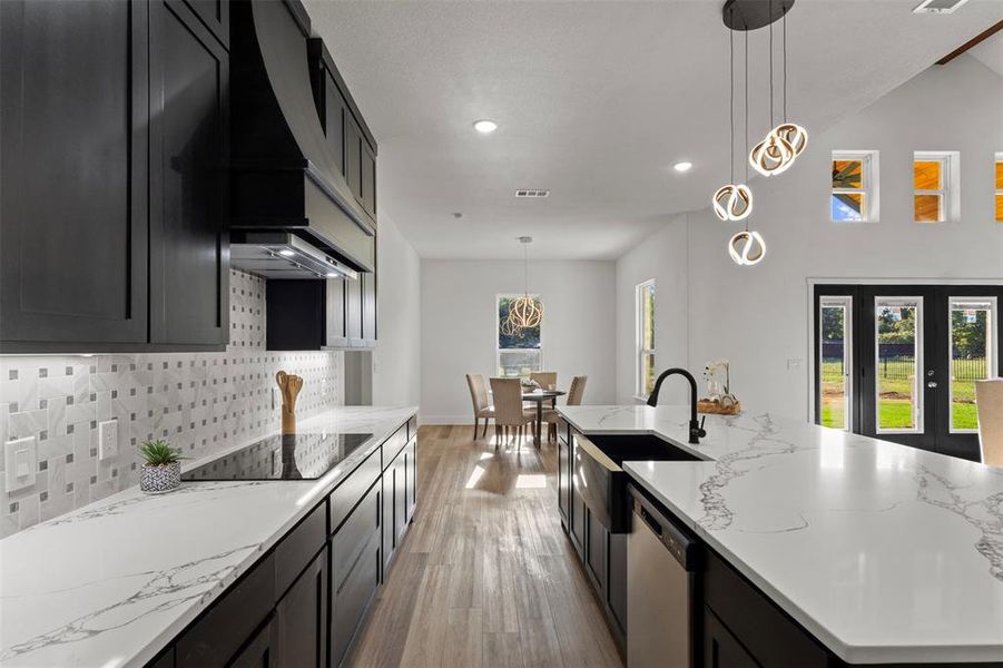 Kitchen featuring dark cabinetry, decorative light fixtures, a large island with sink, light stone countertops, and light wood-style floors