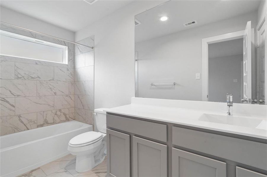 Secondary bath features tile flooring, bath/shower combo with tile surround, stained wood cabinets, beautiful light countertops, mirror, sleek fixtures and modern fini