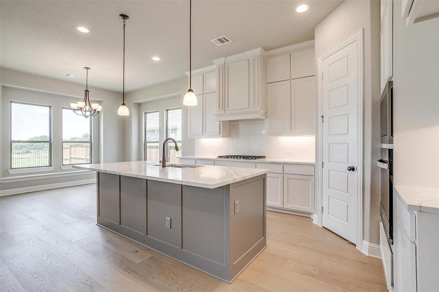 Kitchen featuring light hardwood / wood-style floors, light stone counters, white cabinets, a kitchen island with sink, and a chandelier