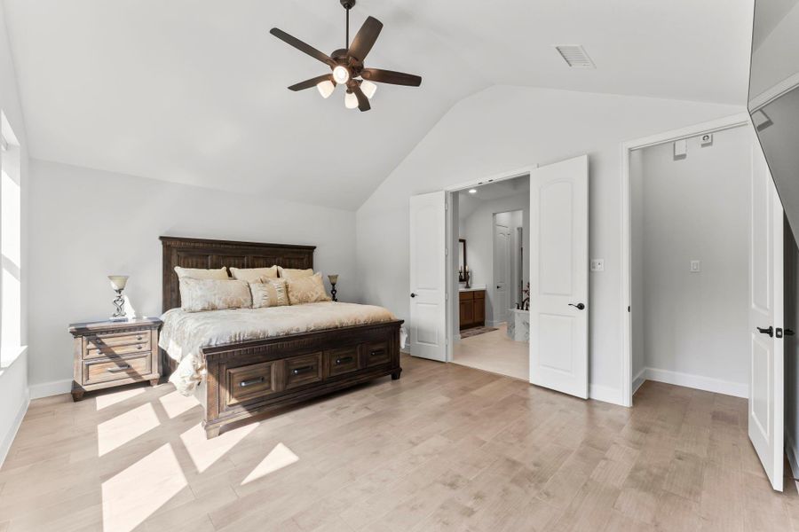 Primary bedroom from an angle that includes the en-suite bathroom. French doors open to reveal the bathroom vanity and toilet. The bedroom has the same dark wood bed and light wood floors. Primary bedroom from an angle that includes the en-suite bathroom. French doors open to reveal the bathroom vanity and toilet. The bedroom has the same dark wood bed and light wood floors.