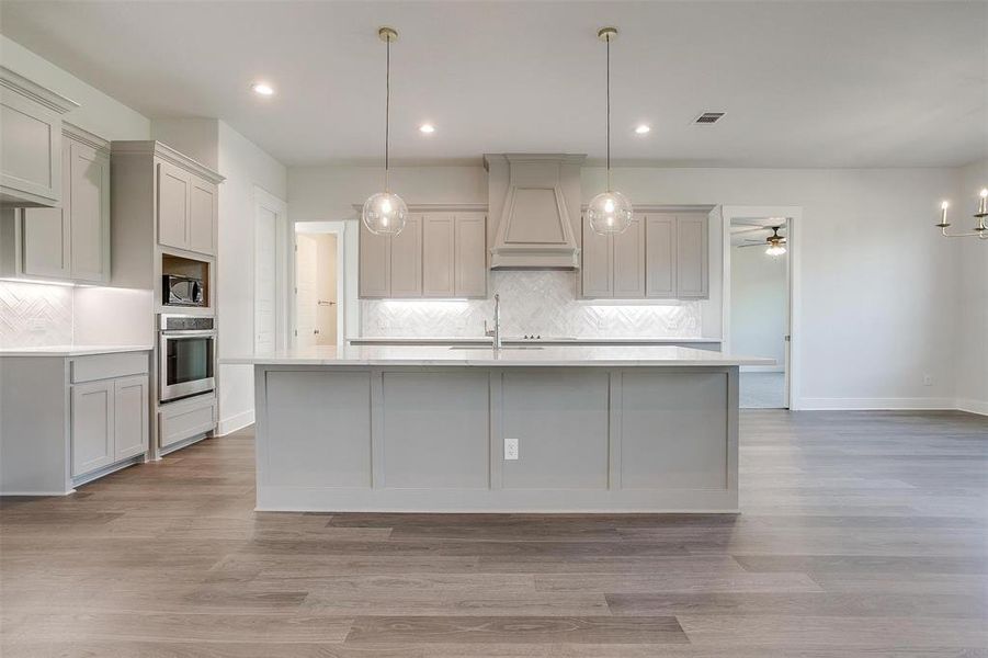 Kitchen featuring backsplash, oven, custom range hood, an island with sink, and light hardwood / wood-style floors