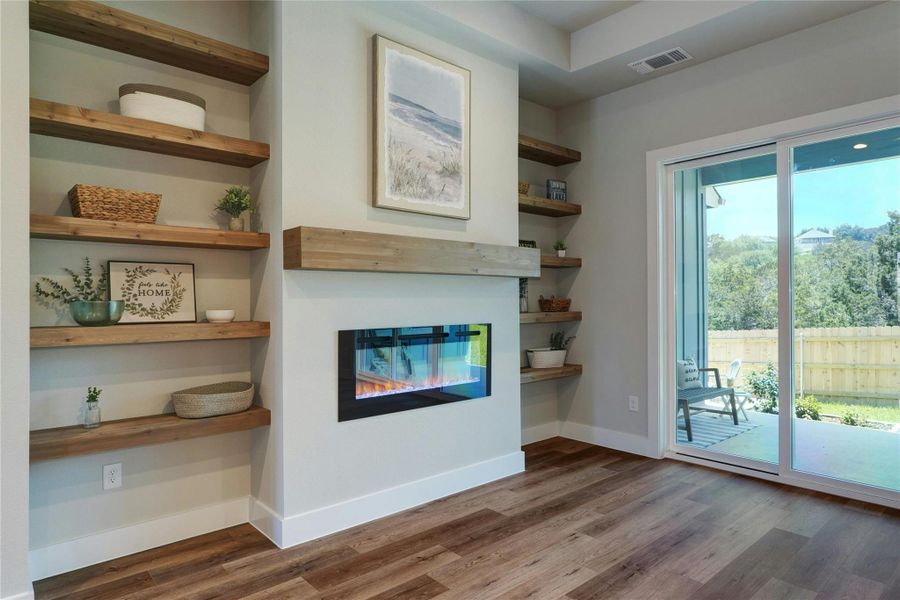 Modern living area featuring a built-in electric fireplace with a wood mantel, integrated shelving, wood-finish flooring, and a sliding glass door opening to an outdoor patio and fenced yard