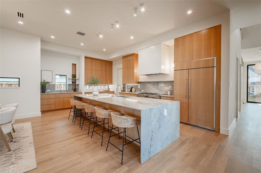 Kitchen featuring light stone counters, a large island with sink, a kitchen breakfast bar, paneled built in fridge, and wall chimney range hood