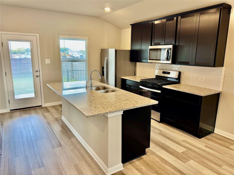 Kitchen featuring stainless steel appliances, decorative backsplash, a center island with sink, light stone countertops, and vaulted ceiling Kitchen featuring stainless steel appliances, decorative backsplash, a center island with sink, light stone countertops, and vaulted ceiling