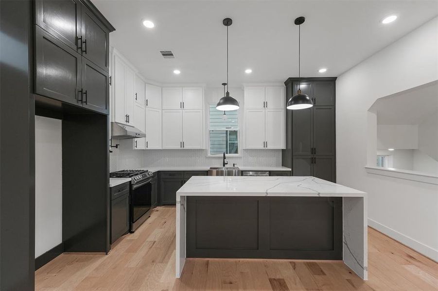 Modern kitchen featuring white countertops, custom dark cabinetry, stainless steel appliances, a central island, and engineered hardwood floors. This is an upgraded kitchen in Plan A.