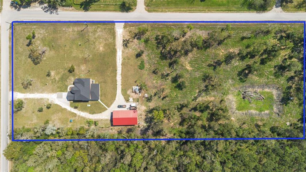 This aerial photo shows a rectangular plot of land with a small house and a red-roofed outbuilding on the left side. The property is bordered by a road at the top and features a mix of open grassy areas and wooded sections, offering potential for development or landscaping.