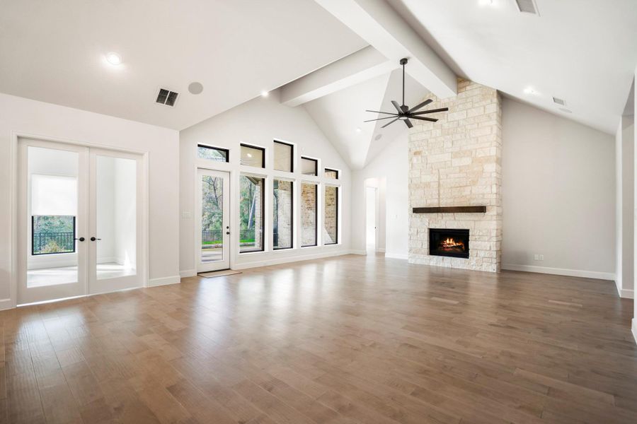 This spacious living room features high vaulted ceilings with exposed beams, a modern ceiling fan, and a cozy stone fireplace. Large windows and glass doors provide plenty of natural light, complemented by warm engineered hardwood floors.