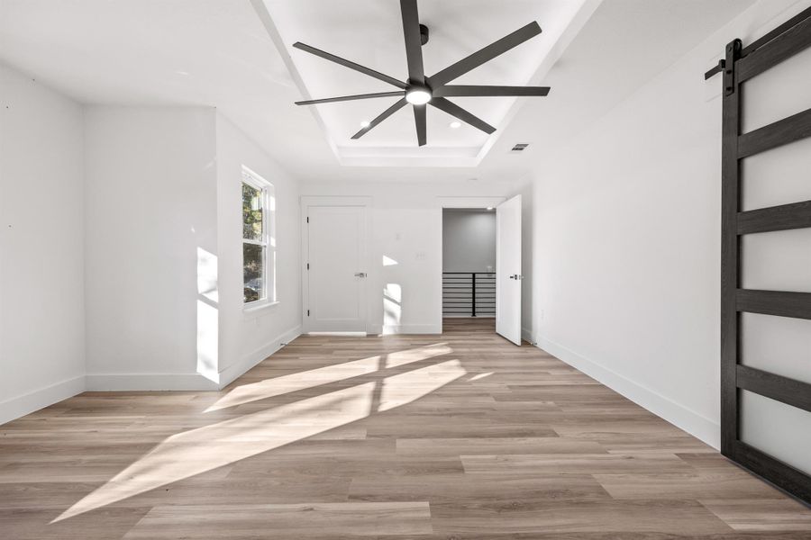 Primary bedroom with a barn door leading to the primary bath, light wood-style flooring, ceiling fan, and a lighted tray ceiling