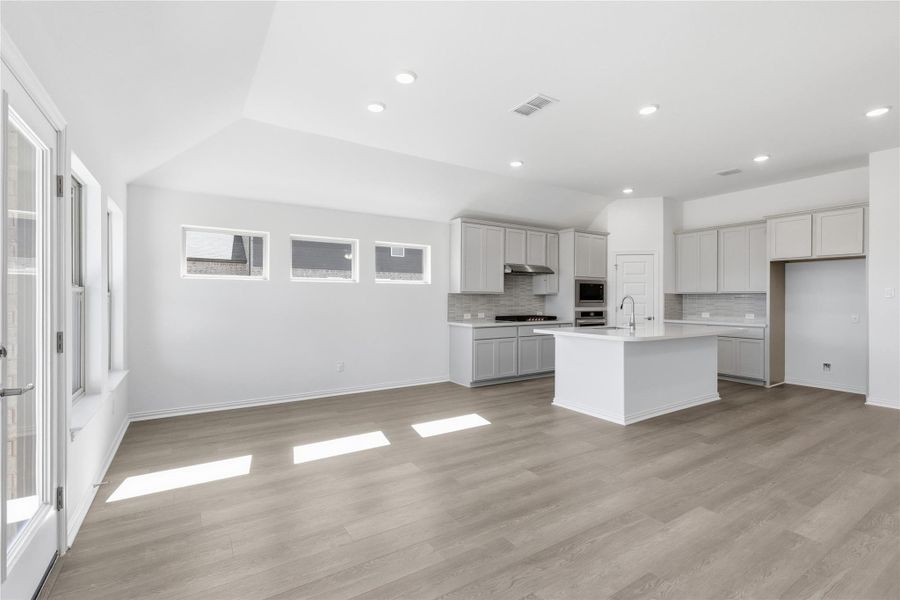Kitchen with a kitchen island with sink, vaulted ceiling, tasteful backsplash, light wood-style floors, and light stone countertops