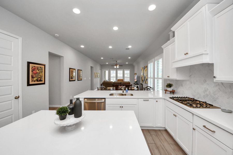 This modern kitchen features sleek white cabinetry, a spacious island with a sink, and a built-in gas cooktop. The open layout leads to a bright living area, enhanced by recessed lighting and large windows, creating an inviting and functional space.