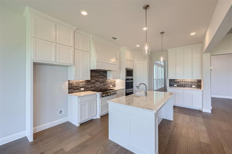 Kitchen with custom range hood, a center island with sink, backsplash, and white cabinetry