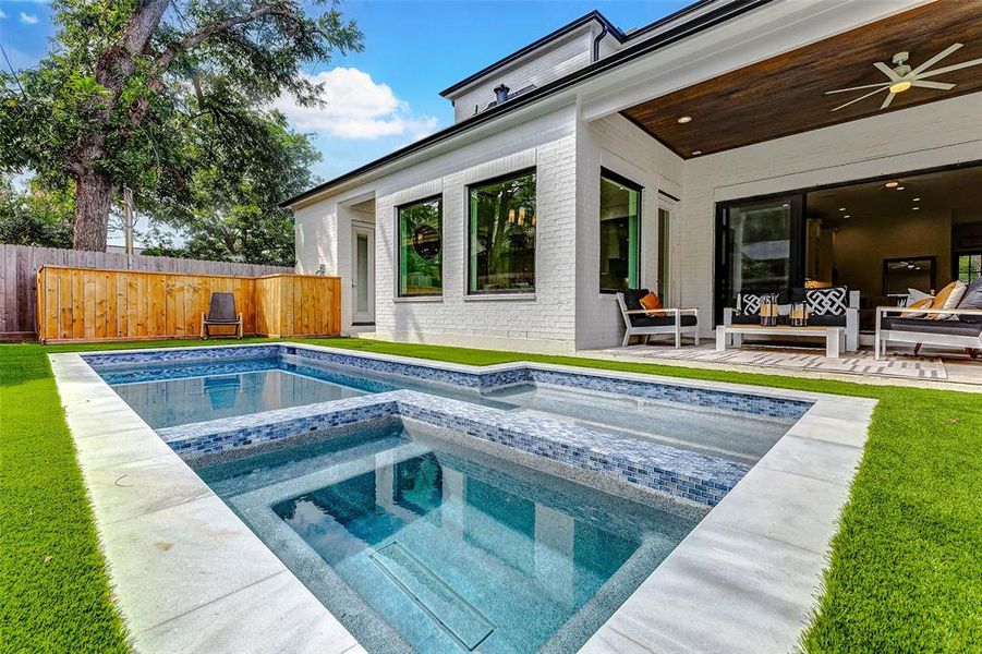 This modern backyard features a luxurious custom-built pool with a spa, surrounded by lush artificial turf. The covered patio includes comfortable seating, perfect for relaxing or entertaining. The serene setting is enhanced by the privacy fence and mature trees.