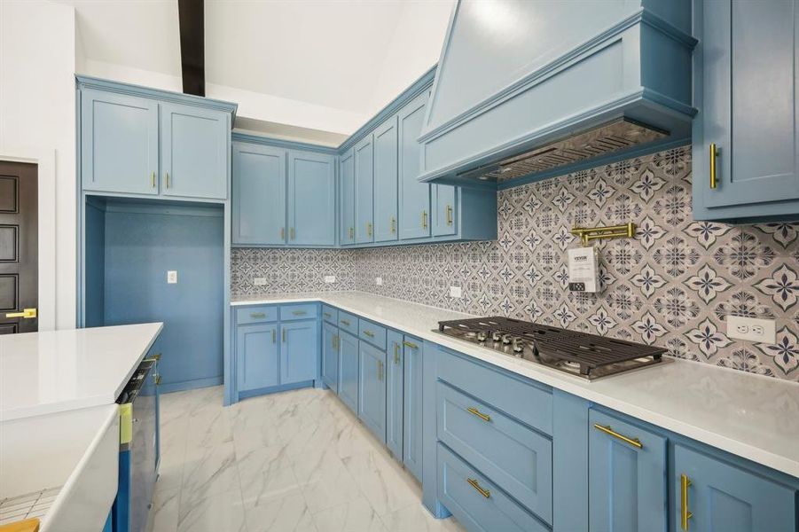 Kitchen featuring tasteful backsplash, custom exhaust hood, light marble finish flooring, stainless steel gas stovetop, and blue cabinets Kitchen featuring tasteful backsplash, custom exhaust hood, light marble finish flooring, stainless steel gas stovetop, and blue cabinets