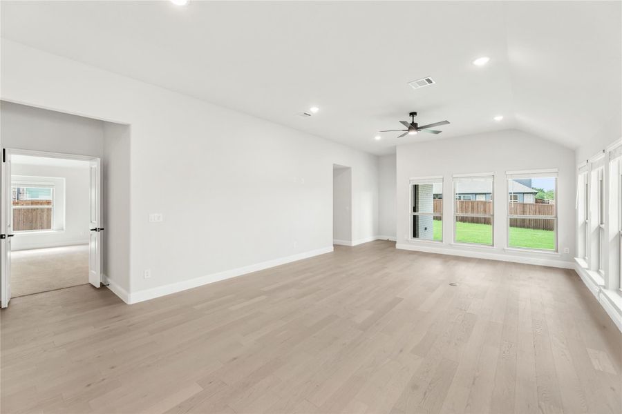 Open living room with light hardwood floors, vaulted ceiling, ceiling fan, and large windows overlooking backyard