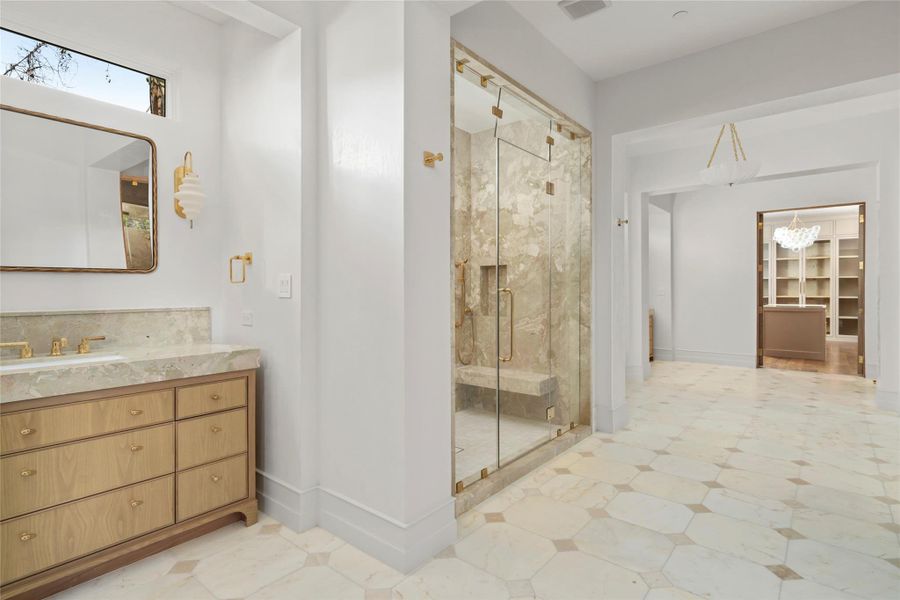 This view of the primary bath highlights its generous proportions and beautifully considered layout, with Botticino tumbled marble flooring extending throughout. A custom vanity, Waterworks fixtures, and an oversized glass-enclosed shower with built-in seating lead seamlessly toward the adjoining custom dressing room. This view of the primary bath highlights its generous proportions and beautifully considered layout, with Botticino tumbled marble flooring extending throughout. A custom vanity, Waterworks fixtures, and an oversized glass-enclosed shower with built-in seating lead seamlessly toward the adjoining custom dressing room.