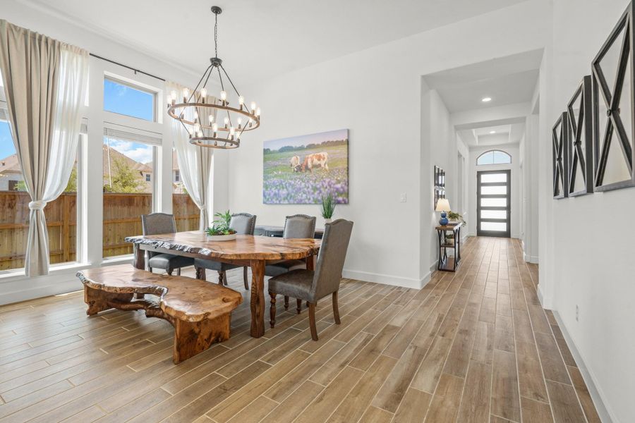 This photo showcases a bright dining area with large windows and elegant curtains. The space has wood-look tile flooring and opens into a welcoming hallway.