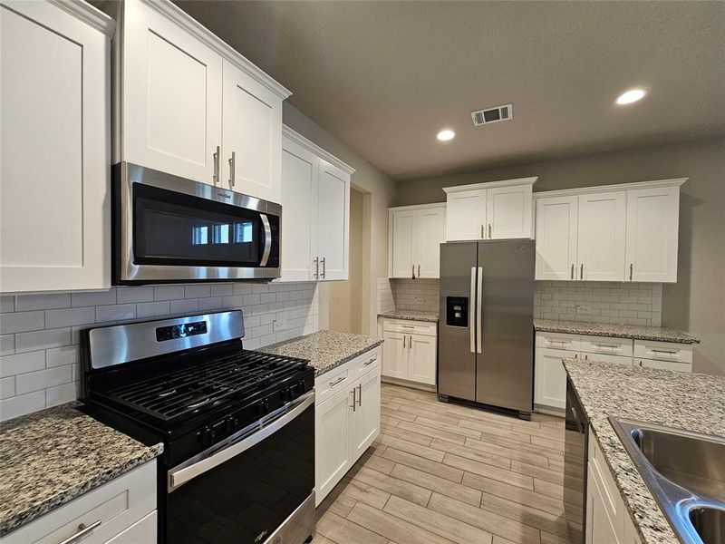 Kitchen with white cabinetry, open floor plan, stainless steel appliances, recess lights, granite counters, backsplash and wood looking tiled floors.