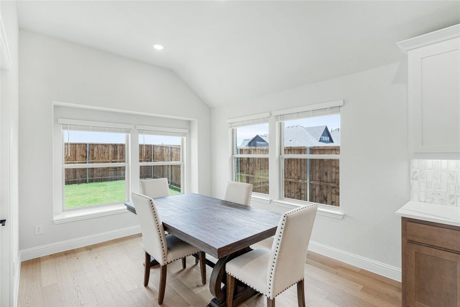 Dining area with dark wood table, four upholstered chairs, vaulted ceiling, and large windows overlooking backyard
