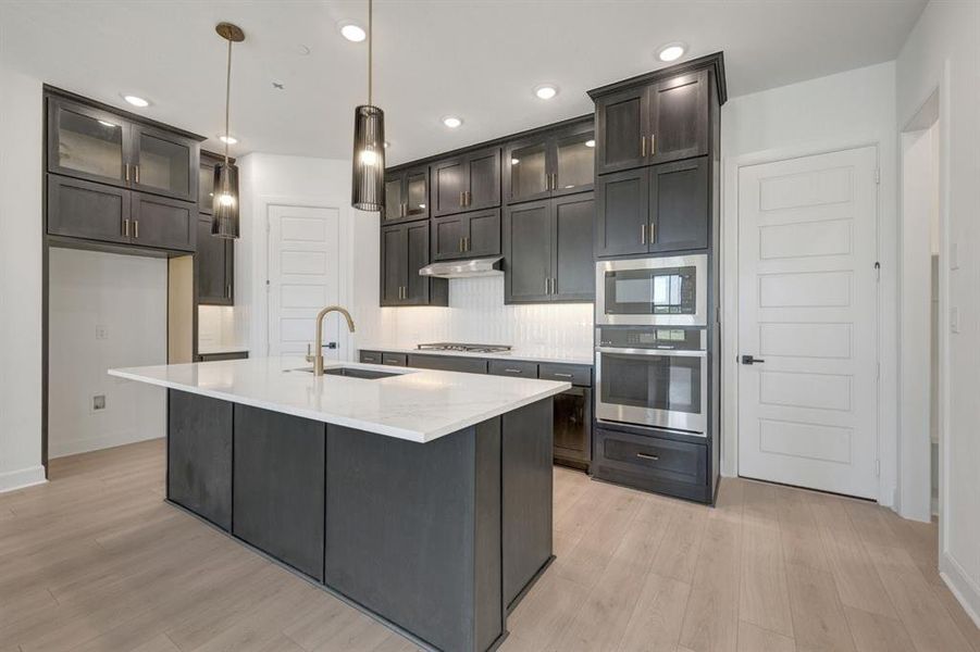 Kitchen featuring a spacious island with a white countertop, dark cabinetry, stainless steel appliances, and wood-finish flooring Kitchen featuring a spacious island with a white countertop, dark cabinetry, stainless steel appliances, and wood-finish flooring