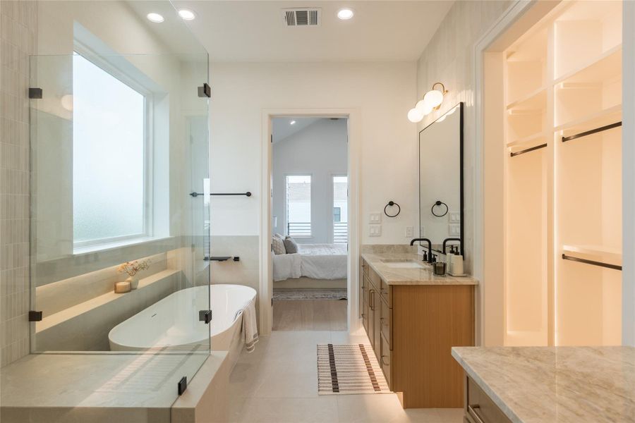 The sight line from the bath back into the primary keeps both spaces in dialogue. A freestanding tub anchors one side, a vanity and illuminated linen tower anchor the other, and through the open doorway the bedroom opens up with its pitched ceiling and balcony sliders beyond. One continuous architectural thought, not two separate rooms.