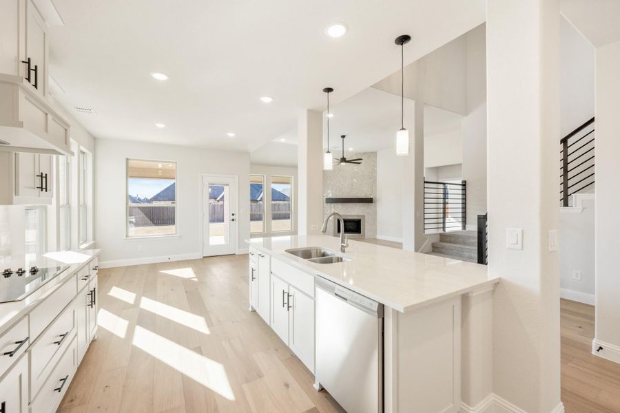 Open-concept kitchen with white cabinets, large island, pendant lights, and light wood floors opening to living area with fireplace.