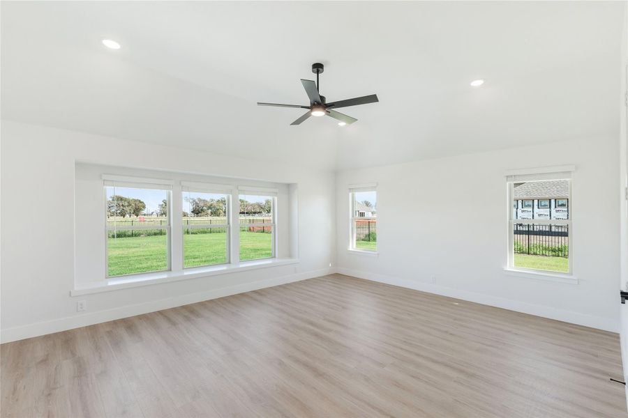 Empty living room with light wood floors, ceiling fan, and large windows overlooking a backyard