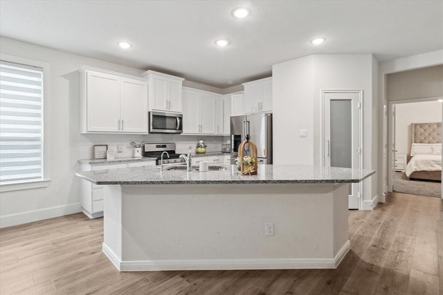 Kitchen featuring white cabinets, light stone counters, light wood-style floors, an island with sink, and stainless steel appliances Kitchen featuring white cabinets, light stone counters, light wood-style floors, an island with sink, and stainless steel appliances