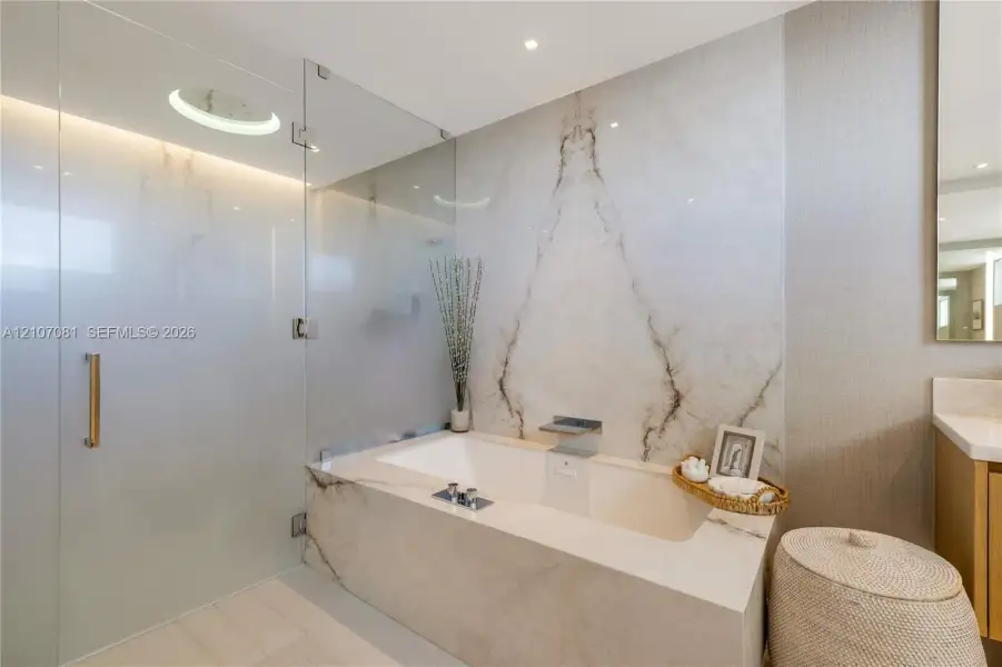 Built-in bathtub featuring full-height natural stone surround, frameless glass shower enclosure, brushed metal fixtures, and recessed ceiling lighting Built-in bathtub featuring full-height natural stone surround, frameless glass shower enclosure, brushed metal fixtures, and recessed ceiling lighting
