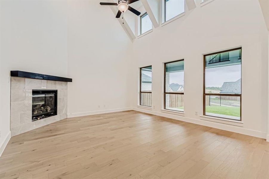 Unfurnished living room featuring light wood-type flooring, a tile fireplace, a towering ceiling, and a ceiling fan Unfurnished living room featuring light wood-type flooring, a tile fireplace, a towering ceiling, and a ceiling fan