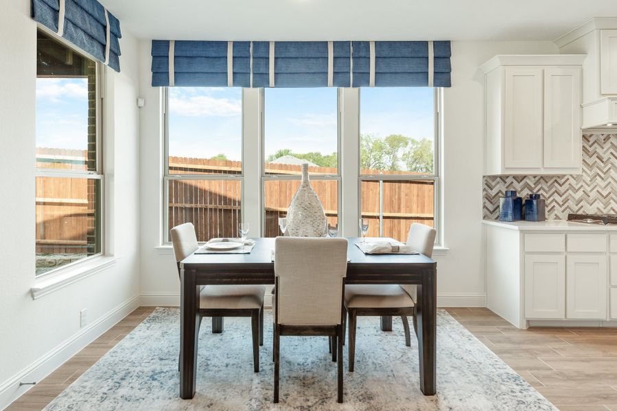Dining area with dark wood table, four chairs, large windows with blue Roman shades, and adjacent white kitchen cabinets