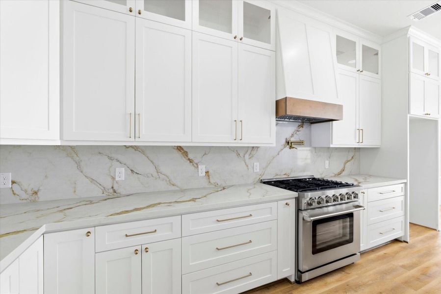 A brightly lit kitchen showcasing a large quartz island, solid wood cabinets with integrated soft-close hinges are visible beneath a large, decorative vent hood. A stainless steel pot filler is mounted over a 36 inch gas range.
