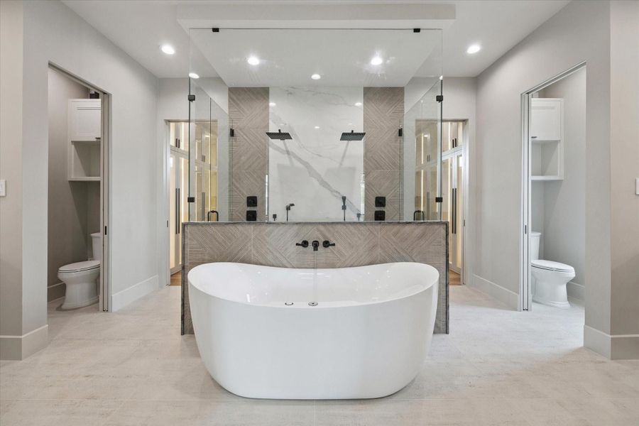 This spa-like primary bathroom is designed to impress with its modern elegance and luxurious finishes. At the center, a freestanding soaking tub creates a striking focal point, perfectly framed by a glass-enclosed shower featuring dual rainfall showerheads and dramatic tile detail.