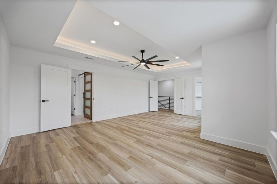 Spacious primary bedroom featuring wood-finish flooring, a tray ceiling with recessed lighting, and a modern ceiling fan Spacious primary bedroom featuring wood-finish flooring, a tray ceiling with recessed lighting, and a modern ceiling fan