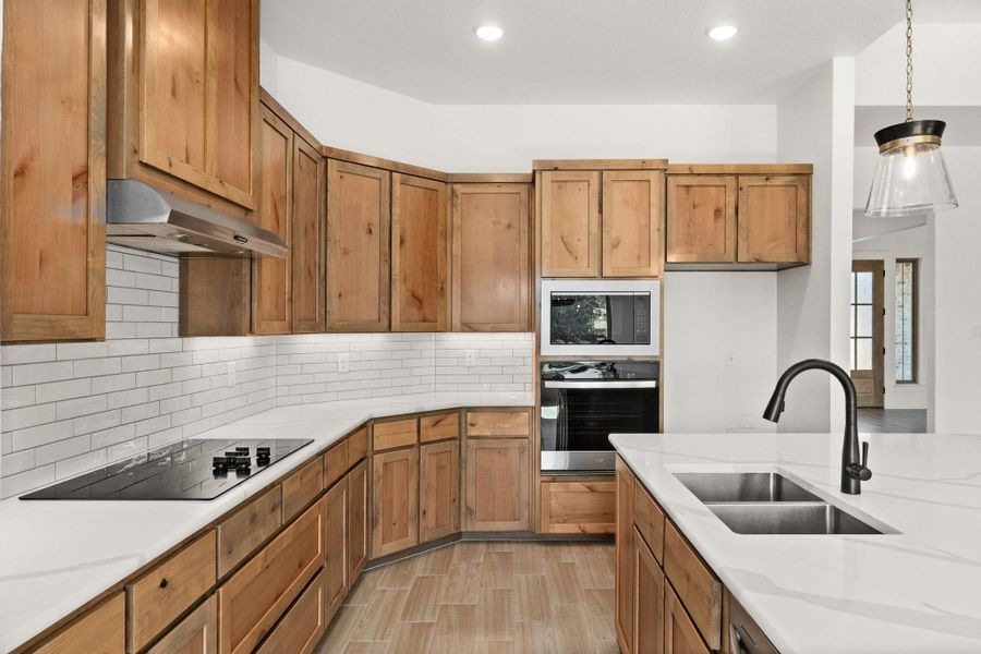 Kitchen featuring brown cabinetry, oven, backsplash, hanging light fixtures, and light stone counters