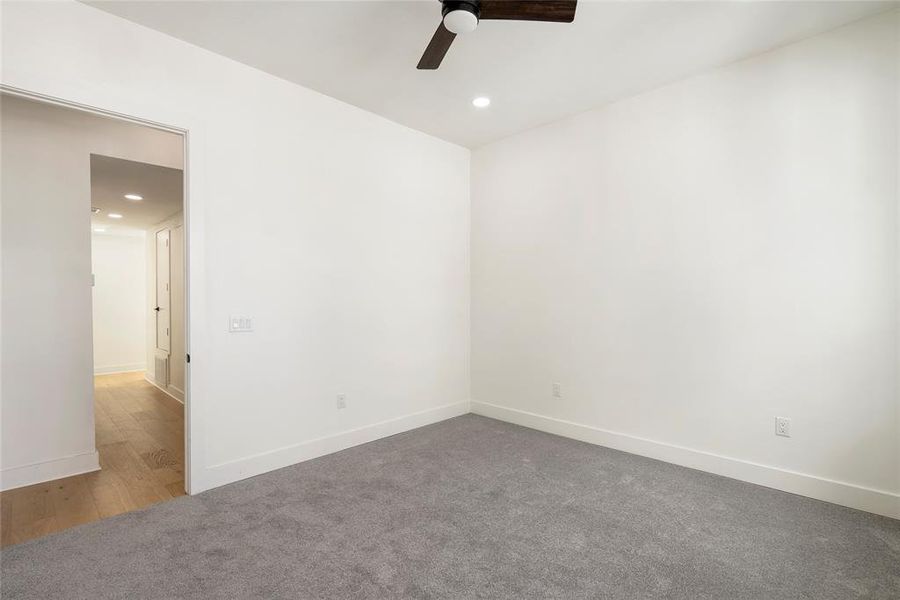 This is a bright and clean room featuring plush gray carpeting, crisp white walls, a dark ceiling fan, and recessed lighting.