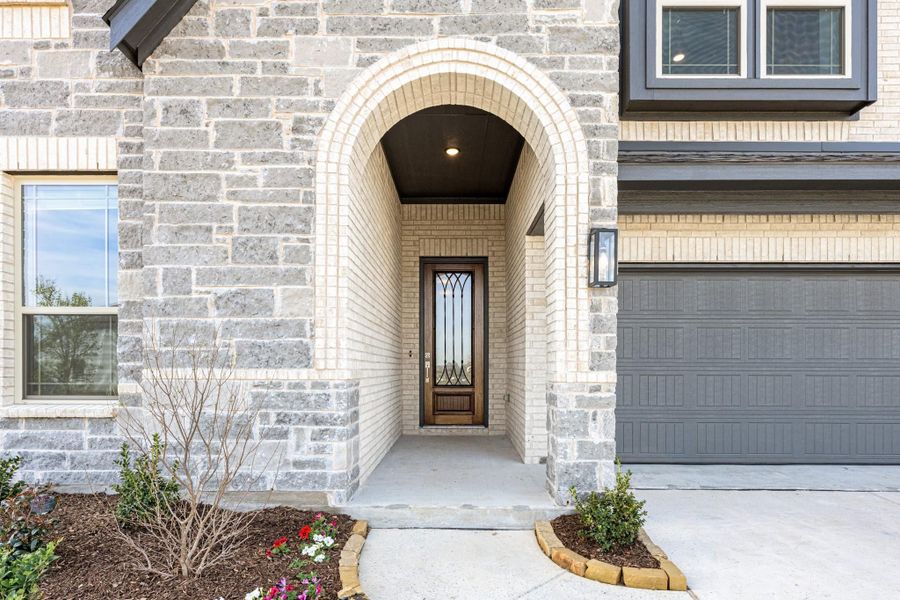Home entrance with arched brick surround, wood and glass front door, and gray garage door beside landscaped walkway. Home entrance with arched brick surround, wood and glass front door, and gray garage door beside landscaped walkway.