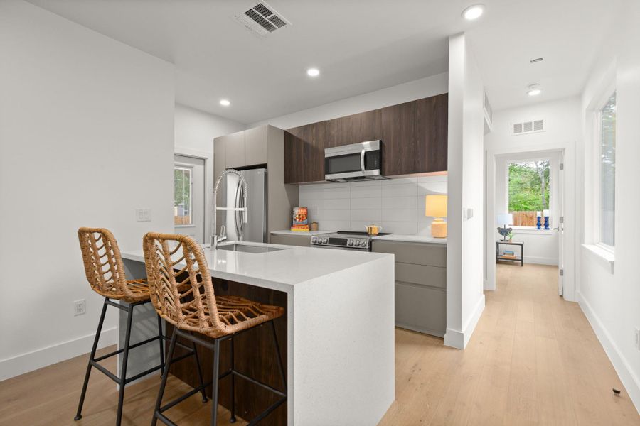 Contemporary kitchen featuring an island with a light-toned countertop, stainless steel appliances, dark wood-finish upper cabinetry, and light gray lower cabinetry