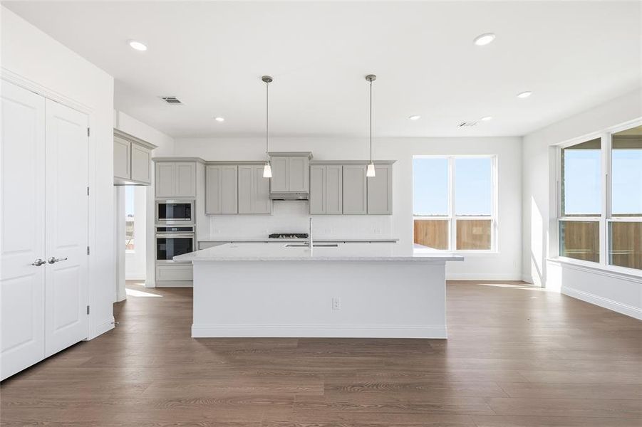 Kitchen featuring gray cabinets, hanging light fixtures, light stone counters, an island with sink, and backsplash Kitchen featuring gray cabinets, hanging light fixtures, light stone counters, an island with sink, and backsplash
