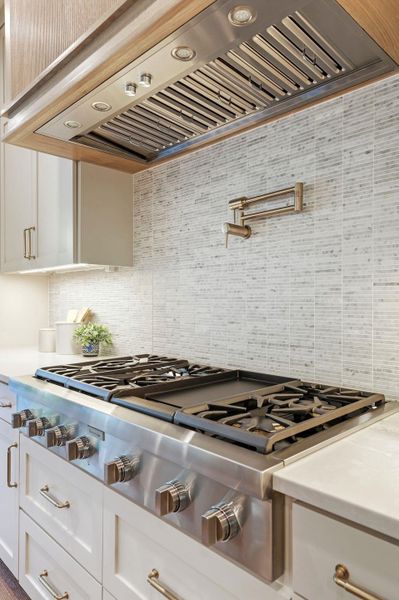 Kitchen with custom exhaust hood, stainless steel gas cooktop, decorative backsplash, light stone countertops, and white cabinetry Kitchen with custom exhaust hood, stainless steel gas cooktop, decorative backsplash, light stone countertops, and white cabinetry