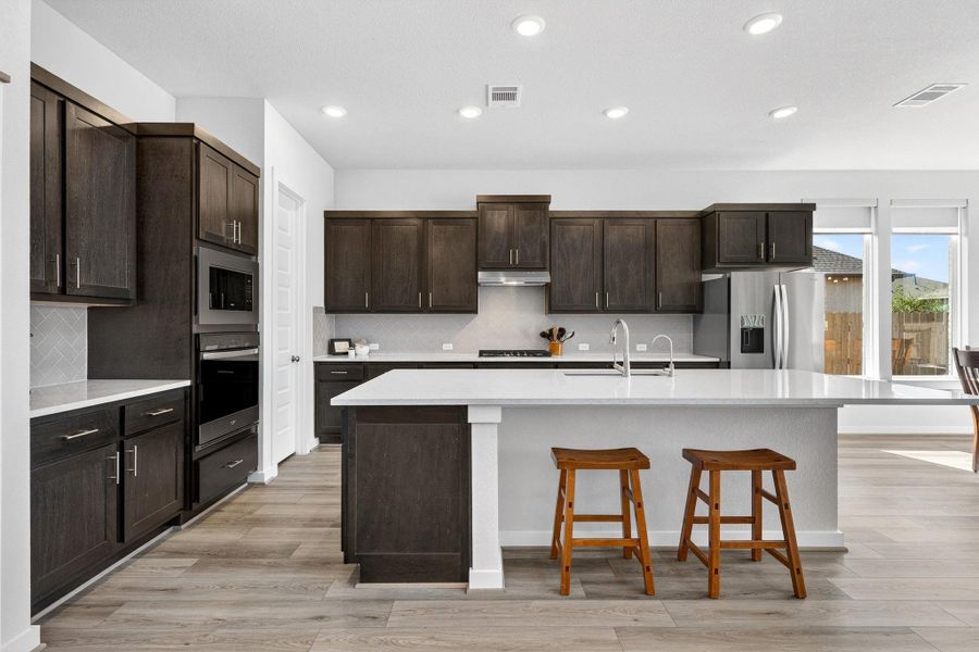 Kitchen with dark wood finish cabinetry,, stainless steel appliances, a built-in oven, a breakfast bar area, and a kitchen island with sink.