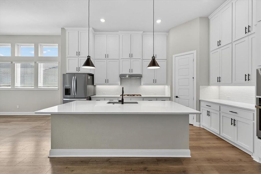 Modern kitchen featuring a large center island with a sink and pendant lighting, extensive white cabinetry with dark hardware, stainless steel appliances, and wood-finish flooring Modern kitchen featuring a large center island with a sink and pendant lighting, extensive white cabinetry with dark hardware, stainless steel appliances, and wood-finish flooring