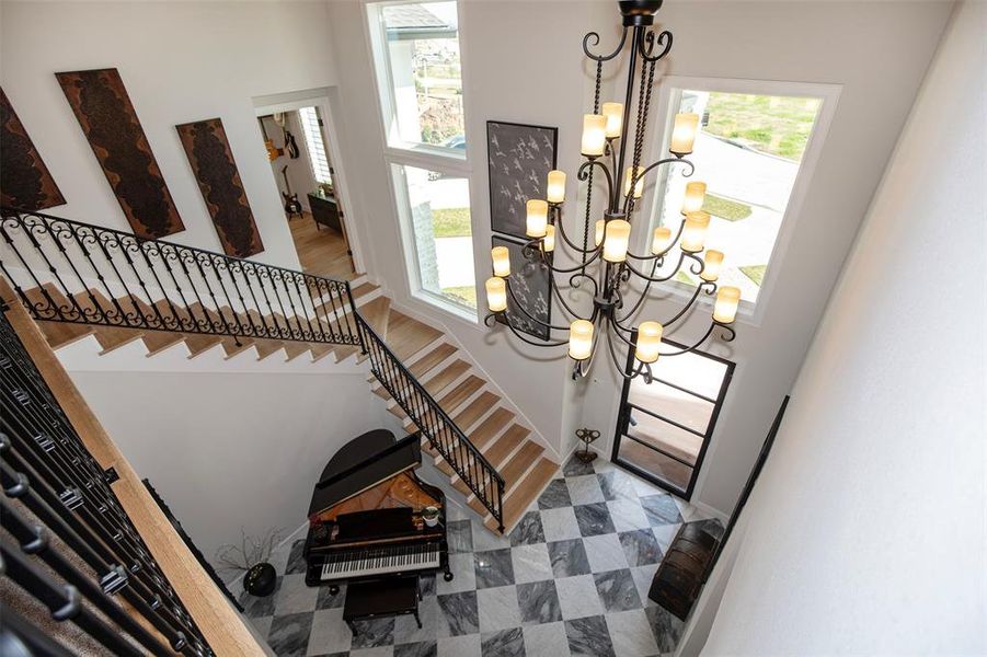 This elegant foyer features a grand staircase with ornate wrought iron railings and a striking chandelier. Large windows provide ample natural light, highlighting the spacious layout and a stylish black and white checkered floor. A grand piano adds a touch of sophistication.