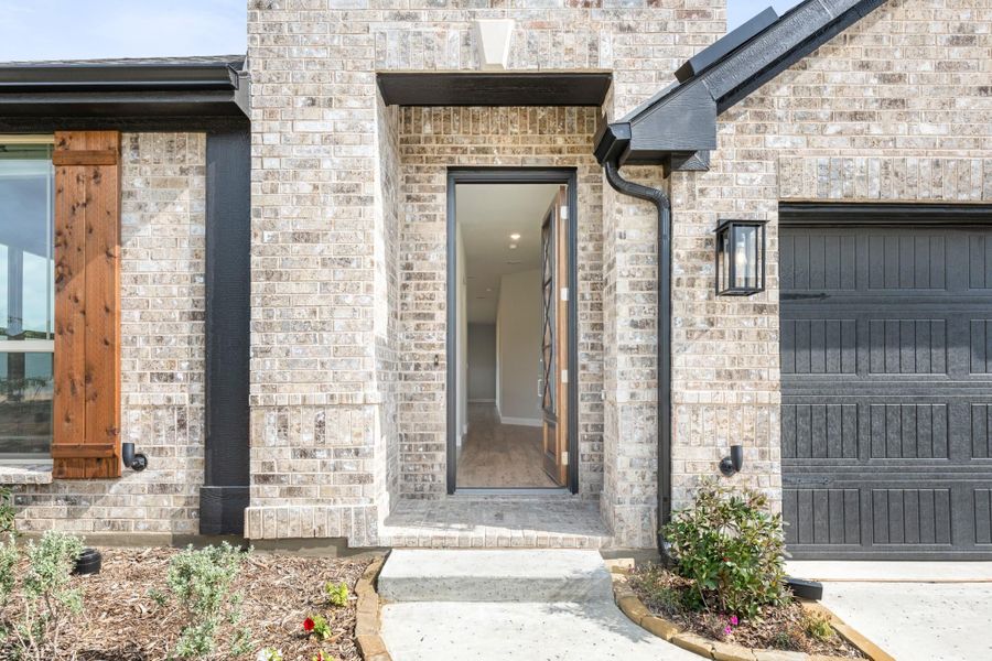 Home entrance with light brick exterior, open wood and glass front door, and dark garage door nearby Home entrance with light brick exterior, open wood and glass front door, and dark garage door nearby