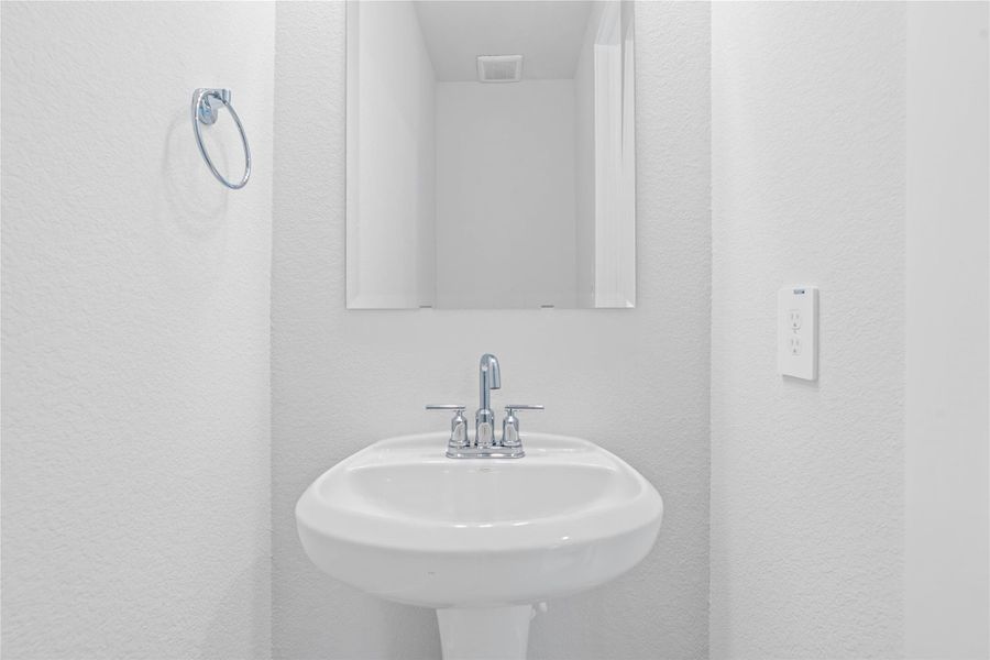 The first-floor powder room offers convenience and a modern aesthetic. It features a classic white pedestal sink complemented by a sleek faucet and a matching wall-mounted towel ring. A simple, large rectangular mirror enhances the sense of space and reflects the bright white walls, making this half-bath both functional and stylish.