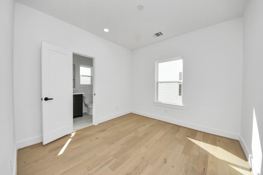This bright, minimalist bedroom features light hardwood floors and a large window that lets in natural light. It includes an en-suite bathroom with modern fixtures, adding convenience and privacy.