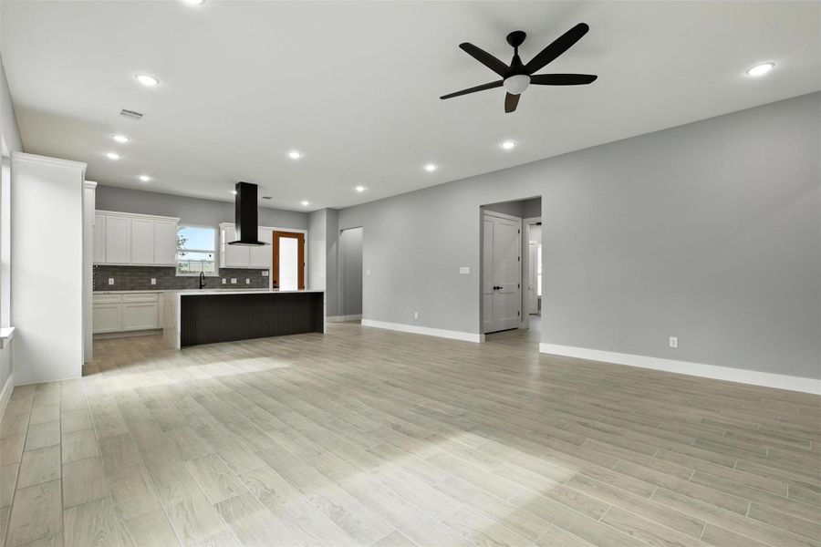 This open-concept living space features light flooring and soft gray walls. The modern kitchen includes white cabinetry, a central island, and stainless steel fixtures. A ceiling fan and recessed lighting provide a comfortable atmosphere.