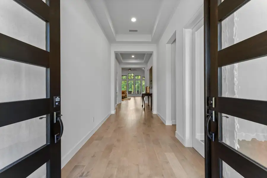 This interior photo shows the home's entryway from just inside the front door. The open front doors have a dark frame with frosted glass panels. The hallway has light wood floors and white walls, leading to a living area with large windows at the back. This interior photo shows the home's entryway from just inside the front door. The open front doors have a dark frame with frosted glass panels. The hallway has light wood floors and white walls, leading to a living area with large windows at the back.