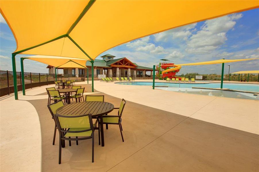 Expansive outdoor patio area featuring multiple shade structures, direct access to a swimming pool with water slide, and a nearby building with stone and wood accents