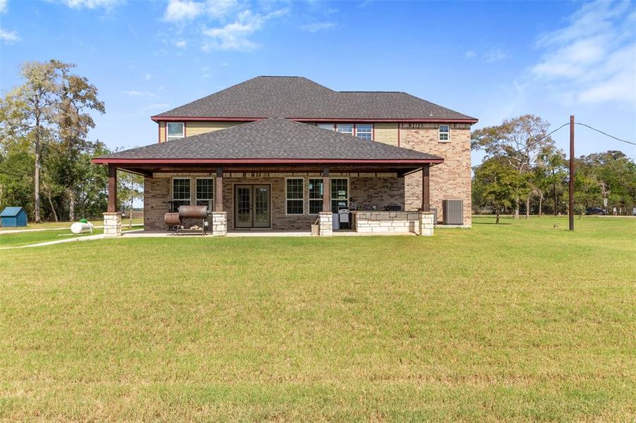 This photo showcases a spacious two-story brick home with a large covered patio, set on a broad, well-maintained lawn. The surrounding area is open and green, providing a serene, rural feel.